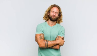 Sarışın adam kollarını kavuşturup mutlu bir şekilde gülüyor, rahat, pozitif ve tatmin edici bir pozla.