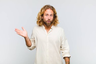 Sarışın adam şaşırmış ve şoka uğramış görünüyor, çenesi düşmüş ve eli açık bir obje tutuyor.