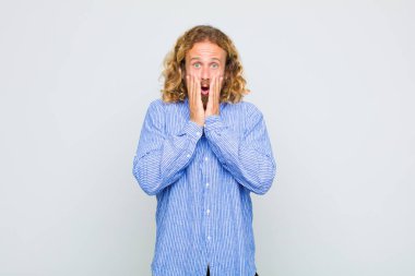 Sarışın adam şok olmuş ve korkmuş hissediyor, ağzı açık ve elleri yanaklarında korkmuş görünüyor.