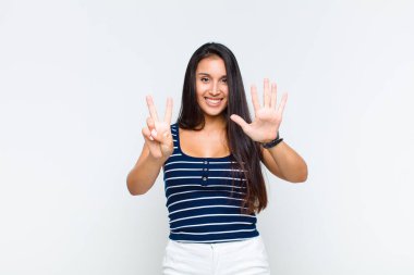Genç bir kadın gülümsüyor ve dost canlısı görünüyor, 7 ya da 7 numarayı gösteriyor, eli önde, geriye doğru sayıyor.
