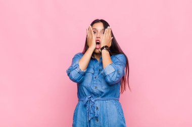 Genç kadın şok olmuş ve korkmuş hissediyor, ağzı açık ve elleri yanaklarında korkmuş görünüyor.