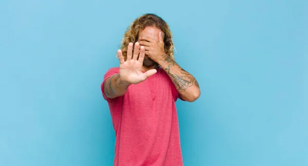 blonde man covering face with hand and putting other hand up front to stop camera, refusing photos or pictures