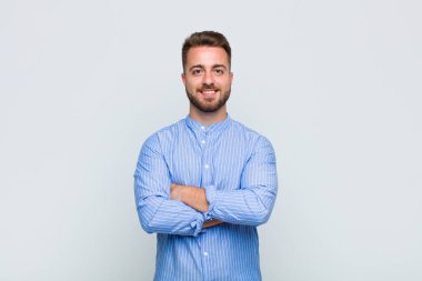 Genç adam kameraya çapraz kollarla gülümsüyor ve mutlu, kendinden emin, tatmin olmuş bir ifade, yan bakış.