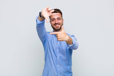 Genç adam mutlu, arkadaş canlısı ve pozitif hissediyor, gülümsüyor ve elleriyle bir portre veya fotoğraf çerçevesi yapıyor.