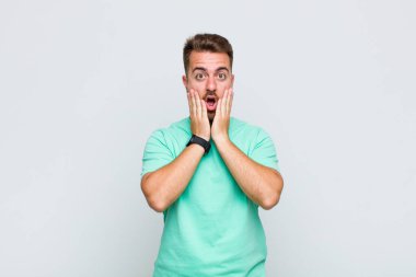 Genç adam şok olmuş ve korkmuş hissediyor, ağzı açık ve elleri yanaklarında korkmuş görünüyor.