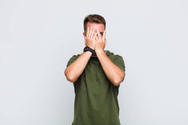 Genç adam elleriyle yüzünü kapatıyor, parmaklarının arasından şaşkın bir ifadeyle yan tarafa bakıyor.
