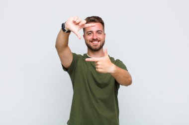 Genç adam mutlu, arkadaş canlısı ve pozitif hissediyor, gülümsüyor ve elleriyle bir portre veya fotoğraf çerçevesi yapıyor.