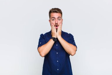 Genç adam şok olmuş ve korkmuş hissediyor, ağzı açık ve elleri yanaklarında korkmuş görünüyor.