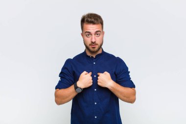 Genç adam şaşkın ve şaşkın bir bakışla kendini işaret ediyor. Seçildiğine şaşırmış ve şaşırmış.