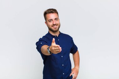 Genç adam gülümsüyor, mutlu görünüyor, kendinden emin ve arkadaş canlısı, bir anlaşma yapmak için el sıkışma teklif ediyor, işbirliği yapıyor.