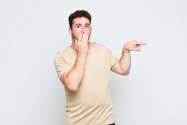 Genç adam mutlu, şaşkın ve şaşırmış hissediyor. Ağzını eliyle kapatıyor ve yanal fotokopi alanını işaret ediyor.