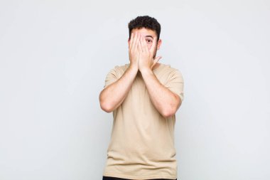 Genç adam elleriyle yüzünü kapatıyor, parmaklarının arasından şaşkın bir ifadeyle yan tarafa bakıyor.
