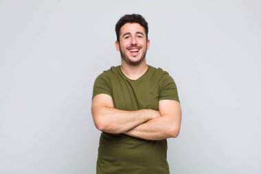 Genç adam mutlu, gururlu ve memnun görünüyor. Kollarını kavuşturup gülümsüyor.