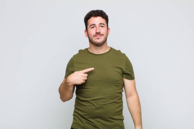 Genç adam gururlu, kendinden emin ve mutlu görünüyor, gülümsüyor ve kendini işaret ediyor veya bir numaralı işareti yapıyor.