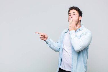 Genç adam mutlu, şaşkın ve şaşırmış hissediyor. Ağzını eliyle kapatıyor ve yanal fotokopi alanını işaret ediyor.