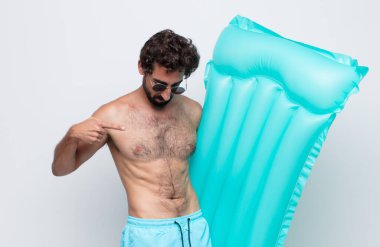 Genç adam neşeyle gülümsüyor, yere bakıyor ve göğsüne işaret ediyor.