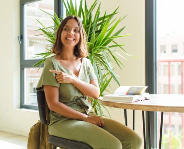 Güzel bir kadın neşeyle gülümsüyor, mutlu hissediyor ve kenarı ve yukarıyı işaret ediyor, fotokopi uzayındaki nesneleri gösteriyor.