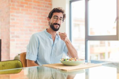 Sakallı bir adam kameraya bakıyor, kendinden emin, arkadaş canlısı bir gülümsemeyle, seni seçiyor.