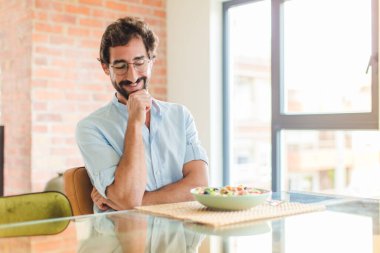 Sakallı bir adam mutlu, kendinden emin bir ifadeyle gülümsüyor, eli çenesinde, merak ediyor ve yan tarafa bakıyor.