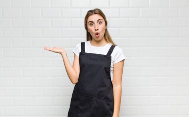 Sarışın kadın şaşırmış ve şoka uğramış görünüyor. Çenesi düşmüş ve eli açık bir objeyi tutuyor.