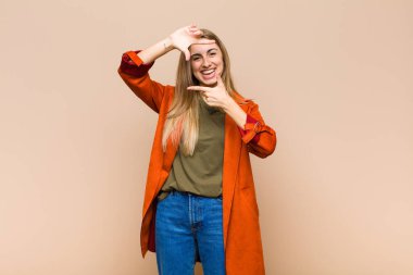 Sarışın kadın mutlu, arkadaş canlısı ve pozitif hissediyor, gülümsüyor ve elleriyle bir portre veya fotoğraf çerçevesi yapıyor.