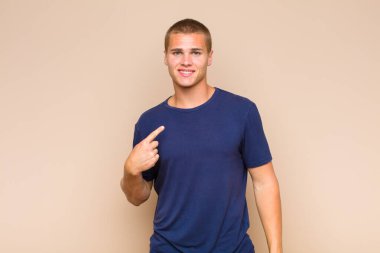 Sarışın adam gururlu, kendinden emin ve mutlu görünüyor, gülümsüyor ve kendini işaret ediyor ya da bir numara işareti yapıyor.