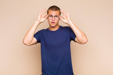 Şok olmuş, şaşırmış ve şaşırmış hisseden sarışın bir adam, elinde şaşkın, inançsız bir bakışla gözlük tutuyor.