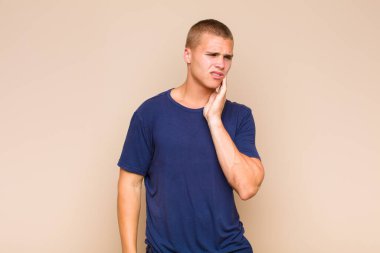 Sarışın adam yanağını tutuyor ve acı çeken diş ağrısı çekiyor, hasta, sefil ve mutsuz hissediyor, dişçi arıyor.