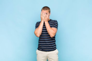 Sarışın adam elleriyle yüzünü kapatıyor, parmaklarının arasından şaşkın bir ifadeyle yan tarafa bakıyor.