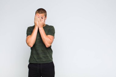 Sarışın adam elleriyle yüzünü kapatıyor, parmaklarının arasından şaşkın bir ifadeyle yan tarafa bakıyor.