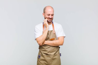 Kel adam yanağını tutuyor ve acı çeken diş ağrısı çekiyor, hasta, sefil ve mutsuz hissediyor, dişçi arıyor.