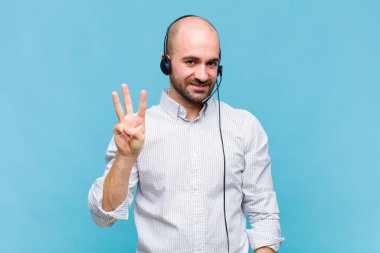 Kel adam gülümsüyor ve arkadaş canlısı görünüyor, üç ya da üç numarayı gösteriyor, eli önde, geriye doğru sayıyor.