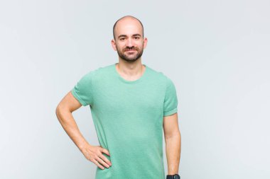 Kel adam mutlu bir şekilde gülümsüyor, eli kalçasında ve kendinden emin, olumlu, gururlu ve arkadaş canlısı.