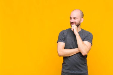 Kel adam mutlu, kendinden emin bir ifadeyle gülümsüyor, çenesinde elleri olan, merak eden ve yan tarafa bakan.