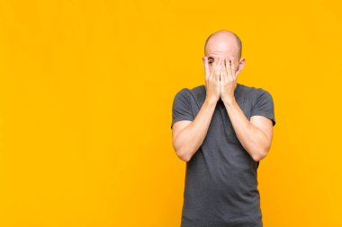 Kel adam elleriyle yüzünü kapatıyor, parmaklarının arasından şaşkın bir ifadeyle yan tarafa bakıyor.