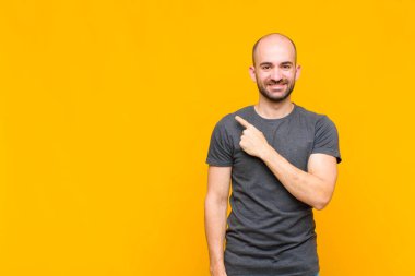 Kel adam neşeyle gülümsüyor, mutlu hissediyor ve kenarı ve yukarıyı işaret ediyor, fotokopi alanında nesneler gösteriyor.