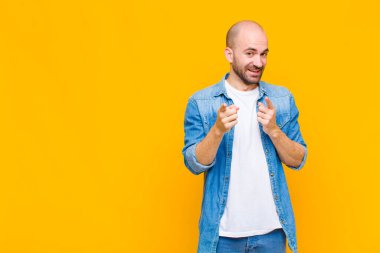 Olumlu, başarılı, mutlu bir tavırla gülümseyen kel bir adam kamerayı işaret ediyor, elleriyle silah işareti yapıyor.