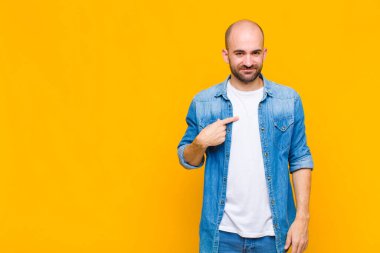 Kel adam gururlu, kendinden emin ve mutlu görünüyor, gülümsüyor ve kendini işaret ediyor ya da bir numara işareti yapıyor.