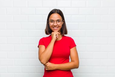 Latin kadın mutlu görünüyor ve gülümsüyor. Elleri çenesinde, merak ediyor ya da soru soruyor. Seçenekleri karşılaştırıyor.