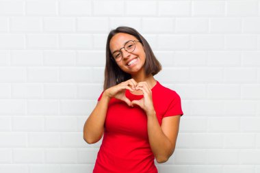 Latin kadın gülümsüyor ve mutlu, sevimli, romantik ve aşık, iki eliyle kalbi şekillendiriyor.