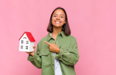Genç İspanyol kadın neşeyle gülümsüyor, mutlu hissediyor ve kenarı ve yukarıyı işaret ediyor, fotokopi alanında nesneler gösteriyor.