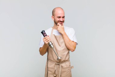 Genç kel bir adam mutlu, kendinden emin bir ifadeyle gülümsüyor, çenesinde elleri, merak ediyor ve yan tarafa bakıyor.