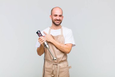 Genç kel adam neşeyle gülümsüyor, mutlu hissediyor ve kenarı ve yukarıyı işaret ediyor, fotokopi alanında nesneler gösteriyor.