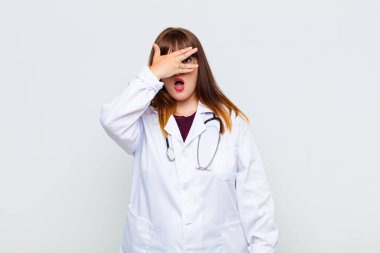 Artı boyunda bir kadın şok olmuş, korkmuş ya da korkmuş görünüyor. Yüzü elle örtülmüş ve parmaklarının arasından gizlice bakıyor.