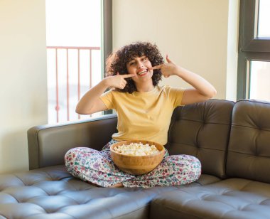 Genç, güzel bir kadın, kendine güvenerek gülümsüyor. Kendi gülüşünü gösteriyor. Olumlu, rahat, tatmin olmuş bir tavır.