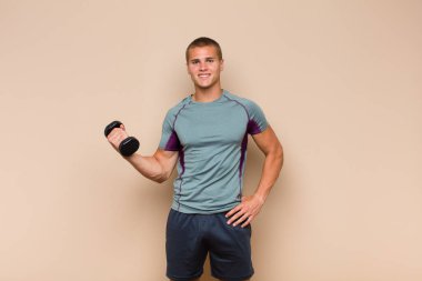 Genç sarışın adam mutlu bir şekilde gülümsüyor, eli kalçasında ve kendinden emin, pozitif, gururlu ve arkadaş canlısı.