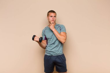 Genç sarışın adam mutlu, kendinden emin bir ifadeyle gülümsüyor. Elleri çenesinde, merak ediyor ve yan tarafa bakıyor.