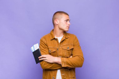 Genç sarışın adam omuz silkiyor, kafası karışık ve kararsız, kolları bağlı ve şaşkın görünüyor.