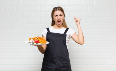 Sarışın, güzel bir kadın sinirli bir ifadeyle bağırıyor ya da yumruklarıyla başarıyı kutluyor.