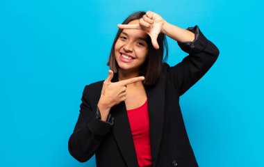 İspanyol kadın mutlu, arkadaş canlısı ve pozitif hissediyor, gülümsüyor ve elleriyle bir portre veya fotoğraf çerçevesi yapıyor.